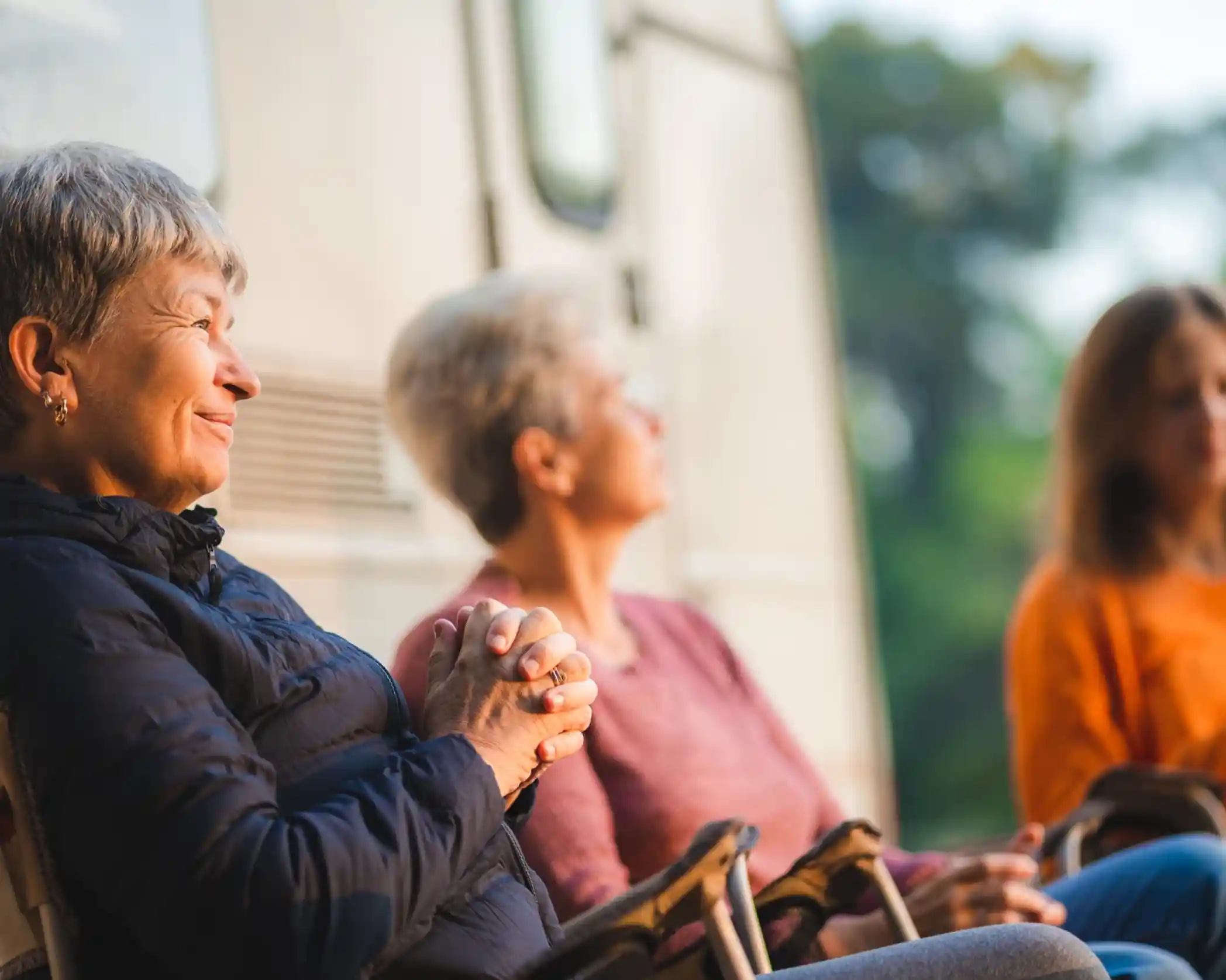Three women relaxing outside, sitting in chairs by a camper van. One woman with short gray hair smiles while the others appear contemplative. Soft lighting and greenery in the background create a serene atmosphere.