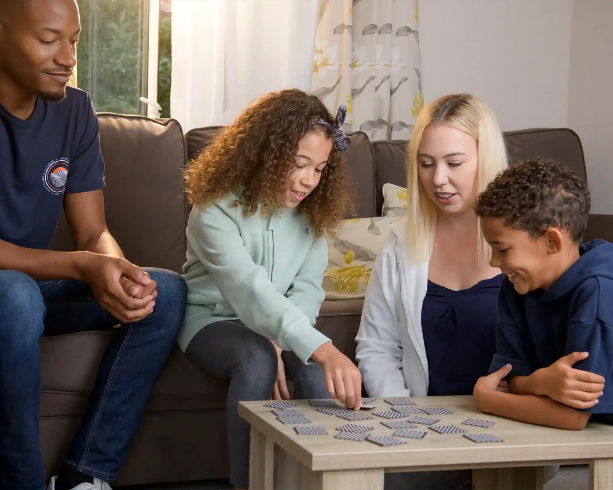A family is seated around a coffee table, playing a memory card game. Two children engage in the game, with one girl pointing at a card while her mother watches. The father smiles from the side, enjoying the moment. The room is bright and cozy.
