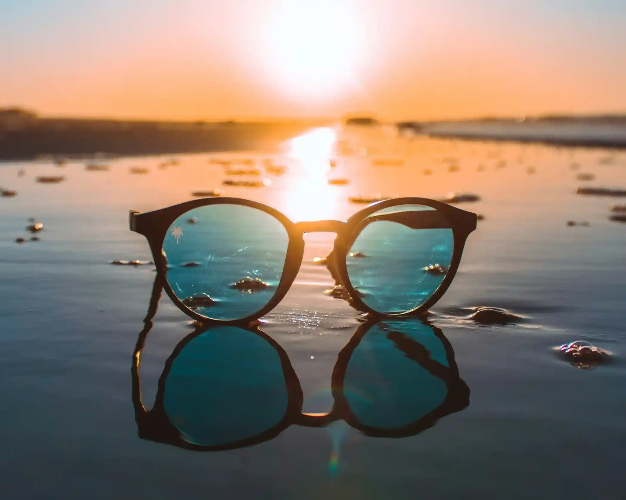 Sunglasses with blue reflective lenses resting on wet sand, with a sunset in the background casting warm tones over the scene.
