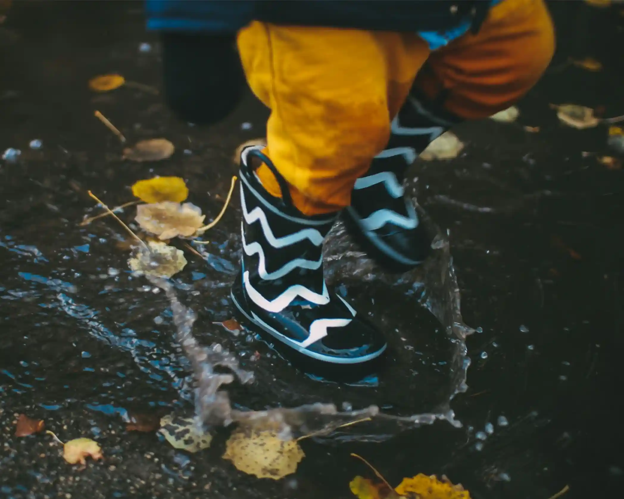 A child in bright orange pants jumps in a puddle while wearing black rain boots with white zig-zag patterns. Autumn leaves are scattered on the wet ground.