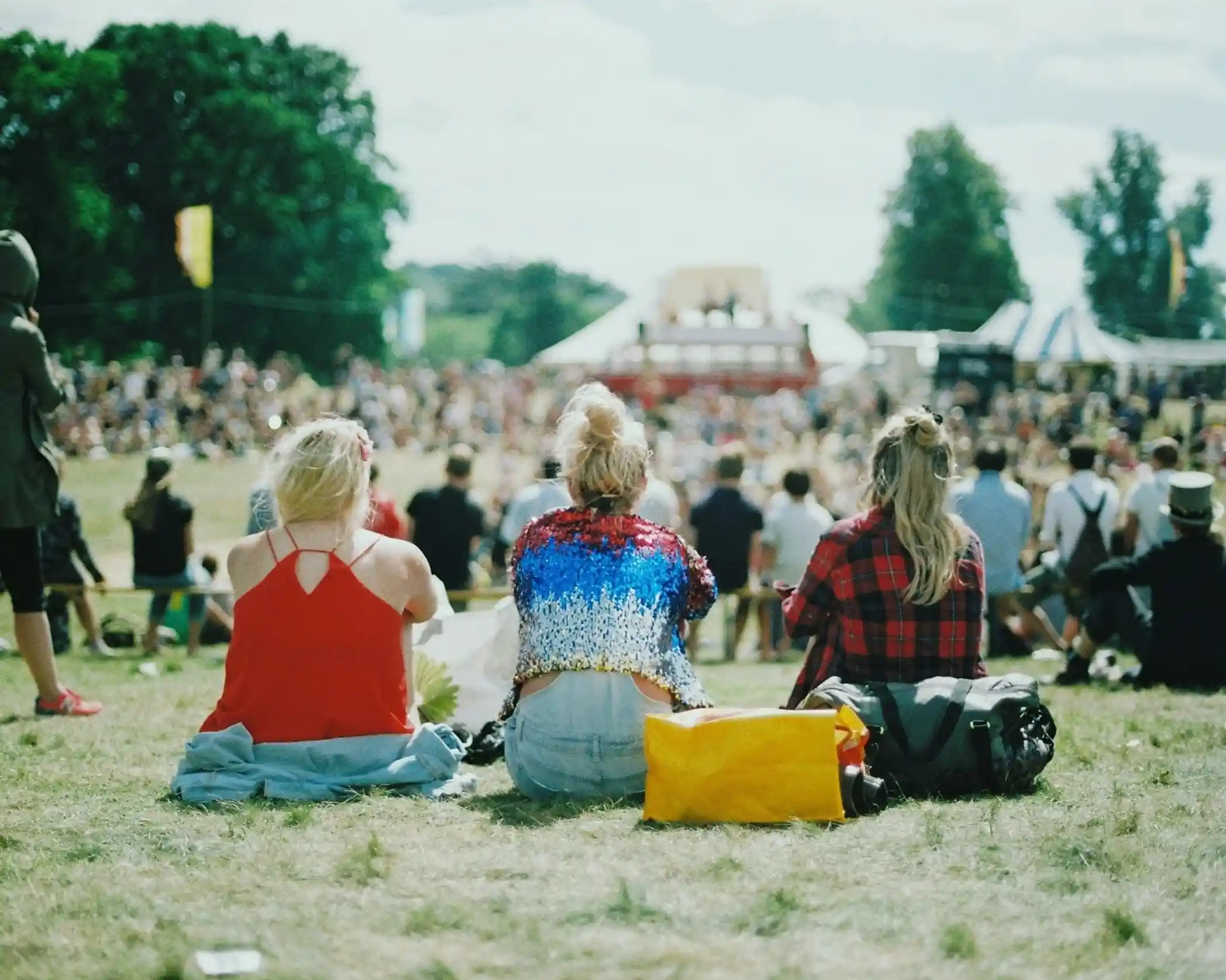 Three women sitting on a grassy hillside, facing a large crowd at an outdoor event. One woman wears a sequined top in red, white, and blue, while the others wear casual clothing. Colorful festival tents and trees are visible in the background.