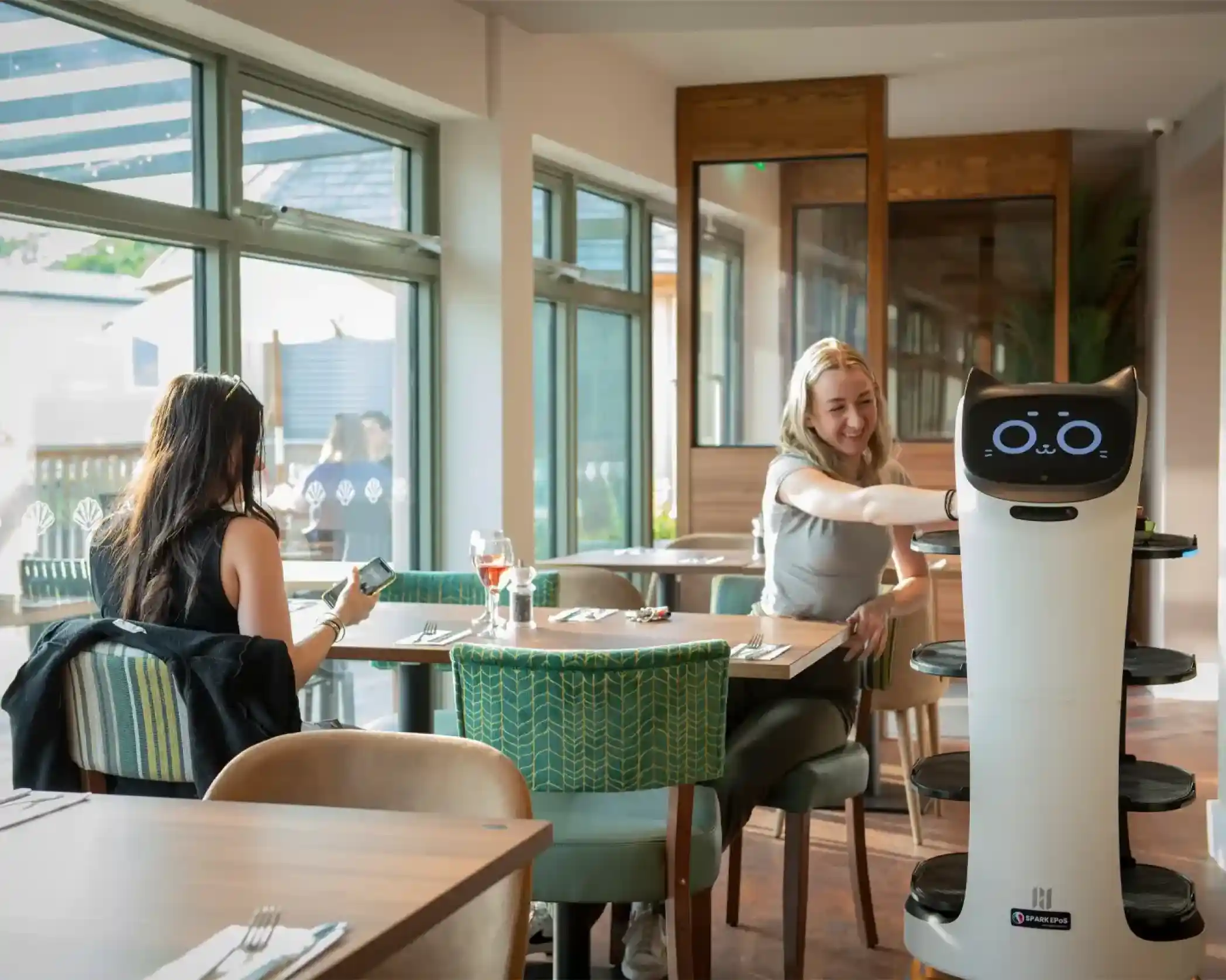 A dining scene in a restaurant featuring a waiter robot serving food to a smiling woman at a table. Another woman is seated nearby, looking at her phone. The interior is bright and modern, with large windows and stylish decor.
