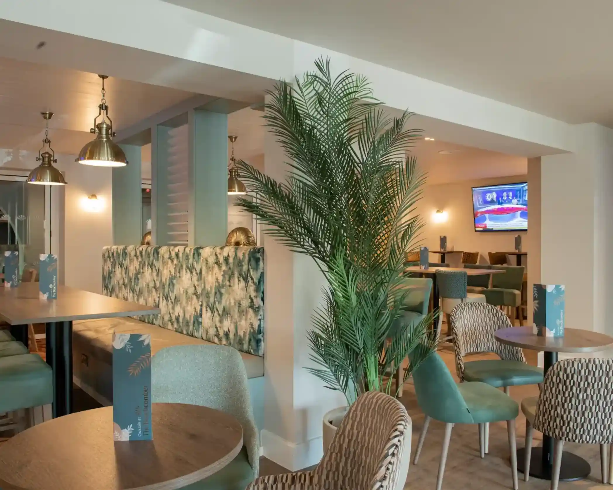 A modern interior of a casual dining area featuring green and teal furniture, decorative palm plants, and soft lighting. There are several tables with stylish chairs, a wall-mounted television, and a cozy seating arrangement in the background.