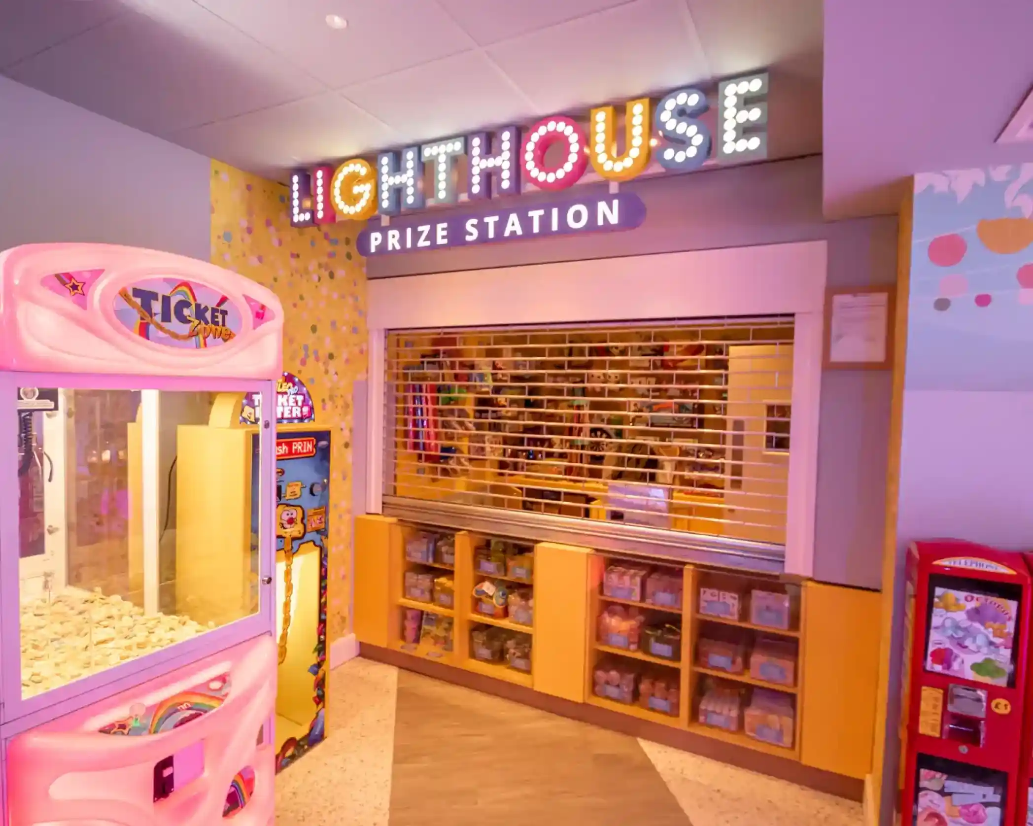 A colorful arcade area featuring the "Lighthouse Prize Station" sign. To the left, a ticket redemption machine and a claw machine are visible. The prize station has a glass window with a counter behind it, showcasing shelves filled with assorted prizes.