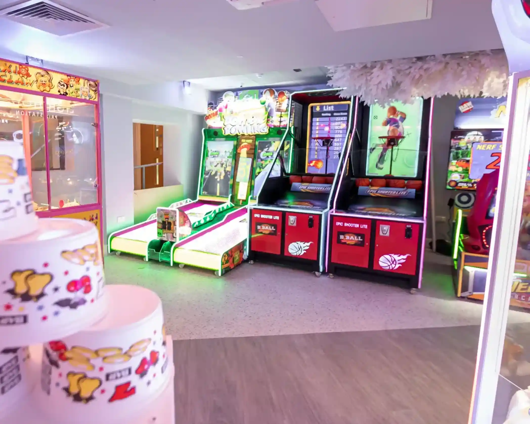 Brightly lit arcade area featuring various games, including a basketball shooting game and a ticket redemption machine. Colorful decorations and playful graphics enhance the atmosphere.
