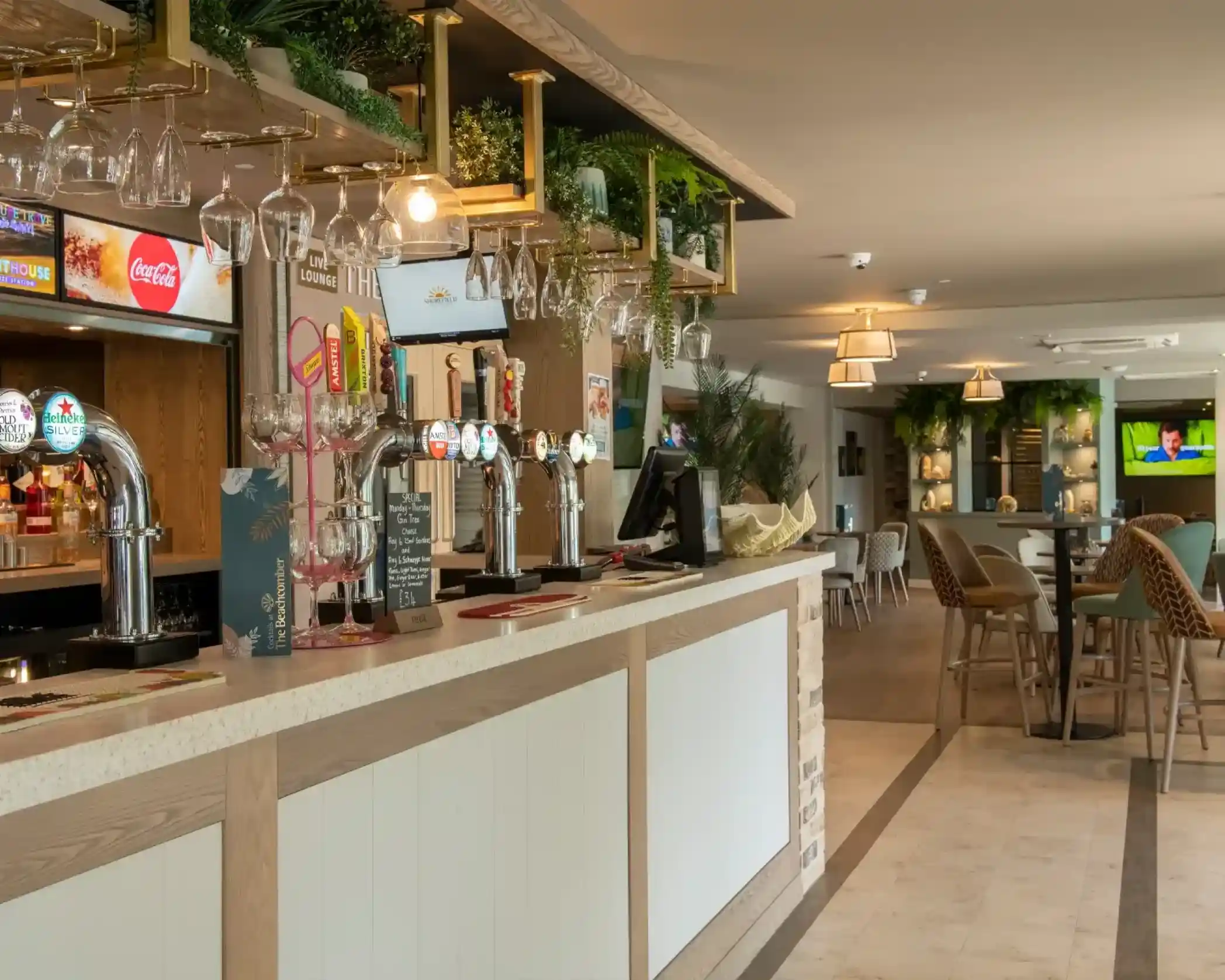 A modern bar area with a wooden counter, various beer taps, and hanging glasses. The background features casual dining tables and chairs, with greenery decoration and a TV displaying a sports game.