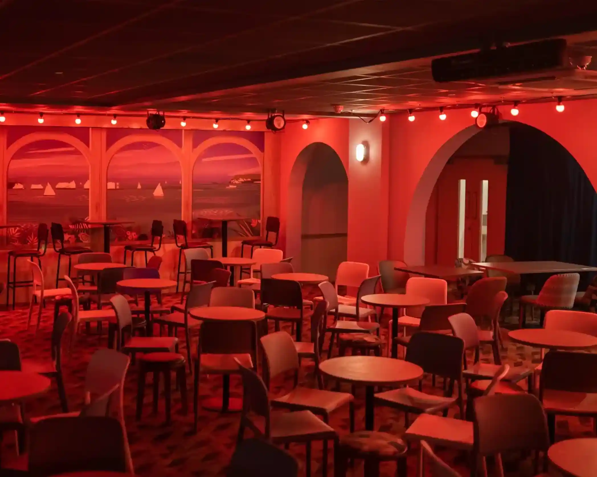 A dimly lit venue with red lighting, featuring numerous empty tables and chairs arranged around the space. Arched windows display a mural of a coastal scene, adding a decorative backdrop.