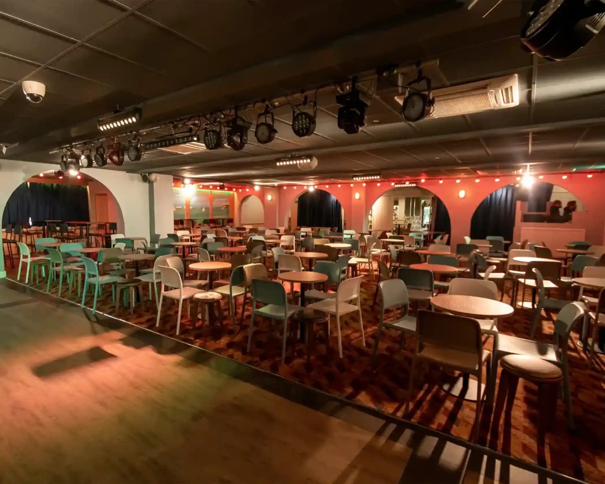 A spacious event venue with rows of tables and chairs arranged in a colorful setting. The walls are adorned with warm lighting and decorative arches, creating an inviting atmosphere.