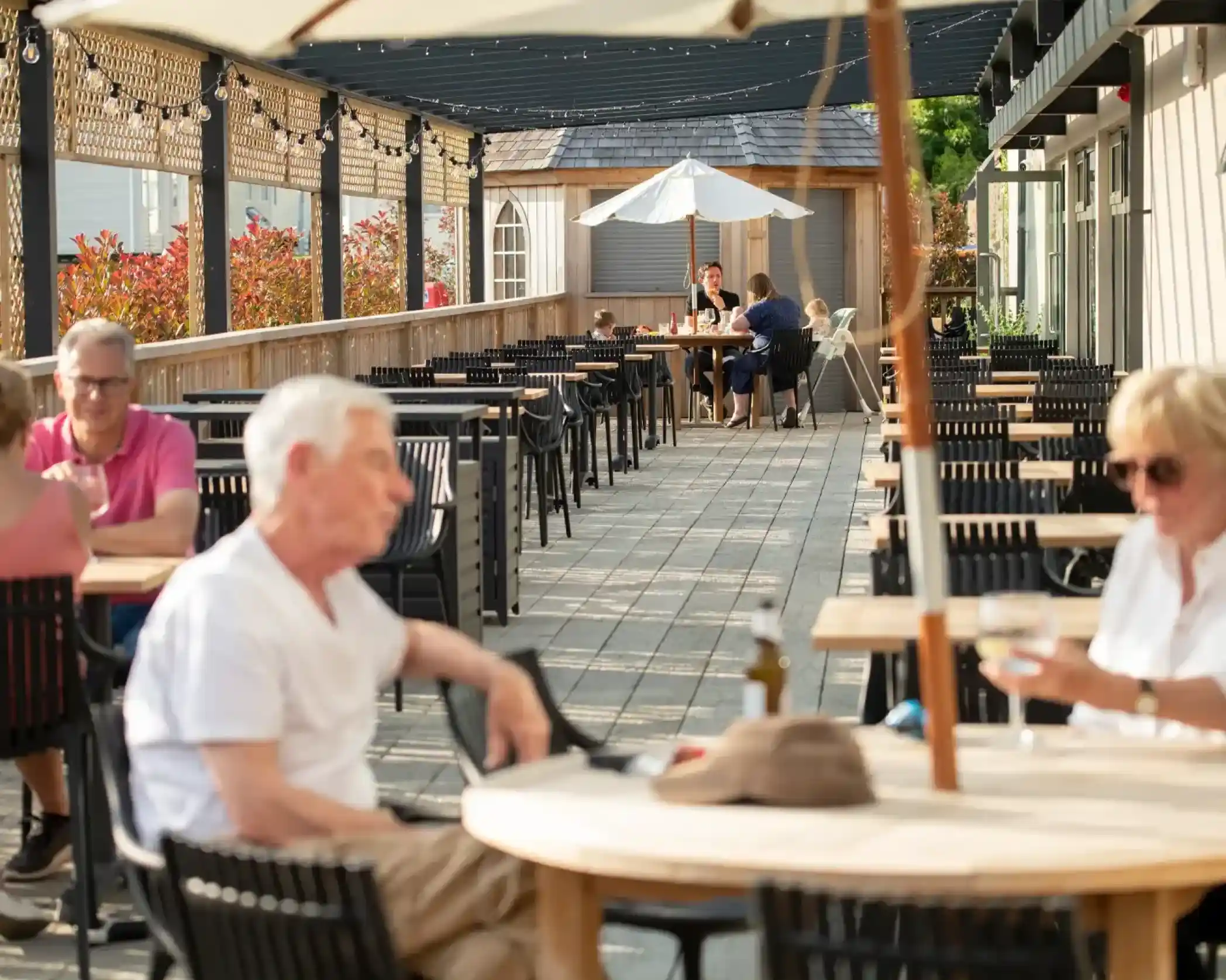 A sunny outdoor terrace at a café, filled with tables and chairs. Several people are seated, some chatting and enjoying drinks. Greenery and colorful flowers line the space, with a large umbrella providing shade.