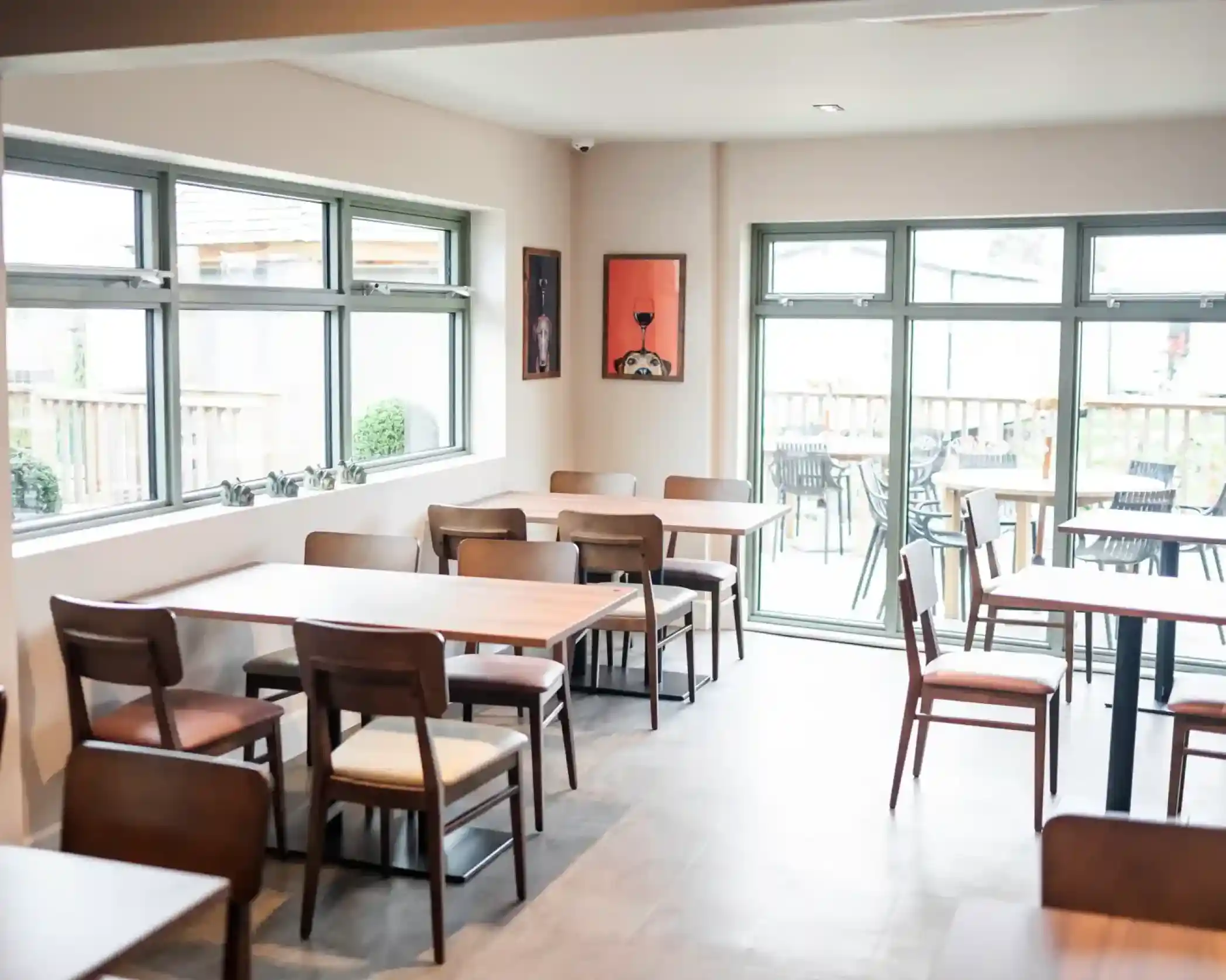 A cozy interior of a café featuring wooden tables and chairs, large windows letting in natural light, and a wall adorned with artwork. Outdoor seating is visible through the glass doors.