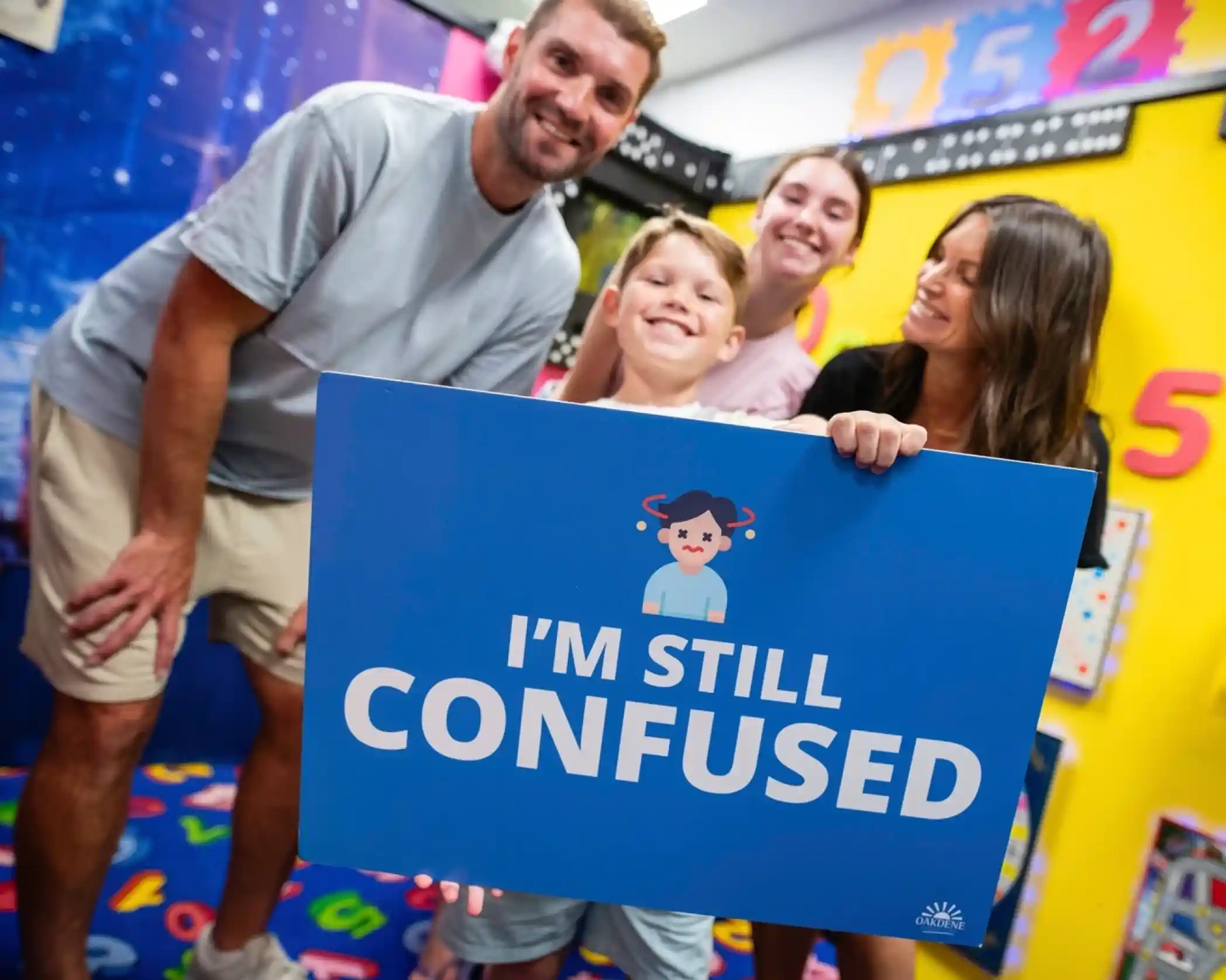 A group of four people poses in a colorful classroom, smiling at the camera. A boy in the front holds a sign that says, "I'm Still Confused," while the adults stand behind him with cheerful expressions.