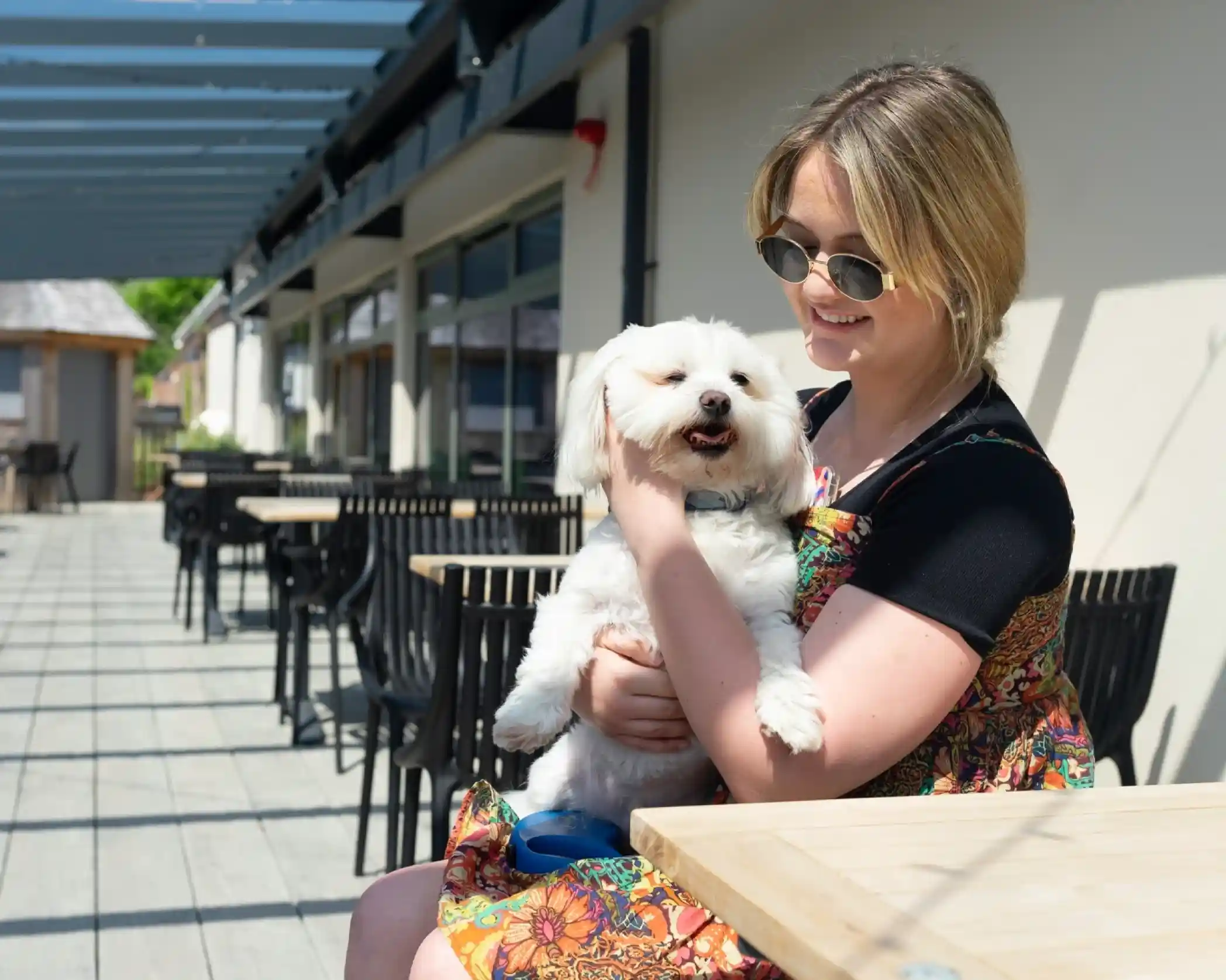 A young woman wearing sunglasses smiles while holding a small white dog in her arms. They are sitting at an outdoor café with wooden tables and chairs in the background.