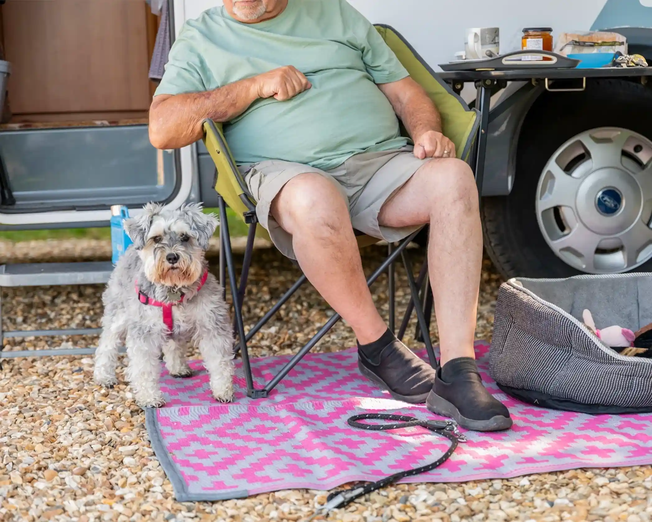 A man seated in a chair at a campsite, wearing a light green shirt and shorts, with a gray schnauzer on a leash beside him. A patterned pink mat lies on the ground, and a camping trailer is in the background.