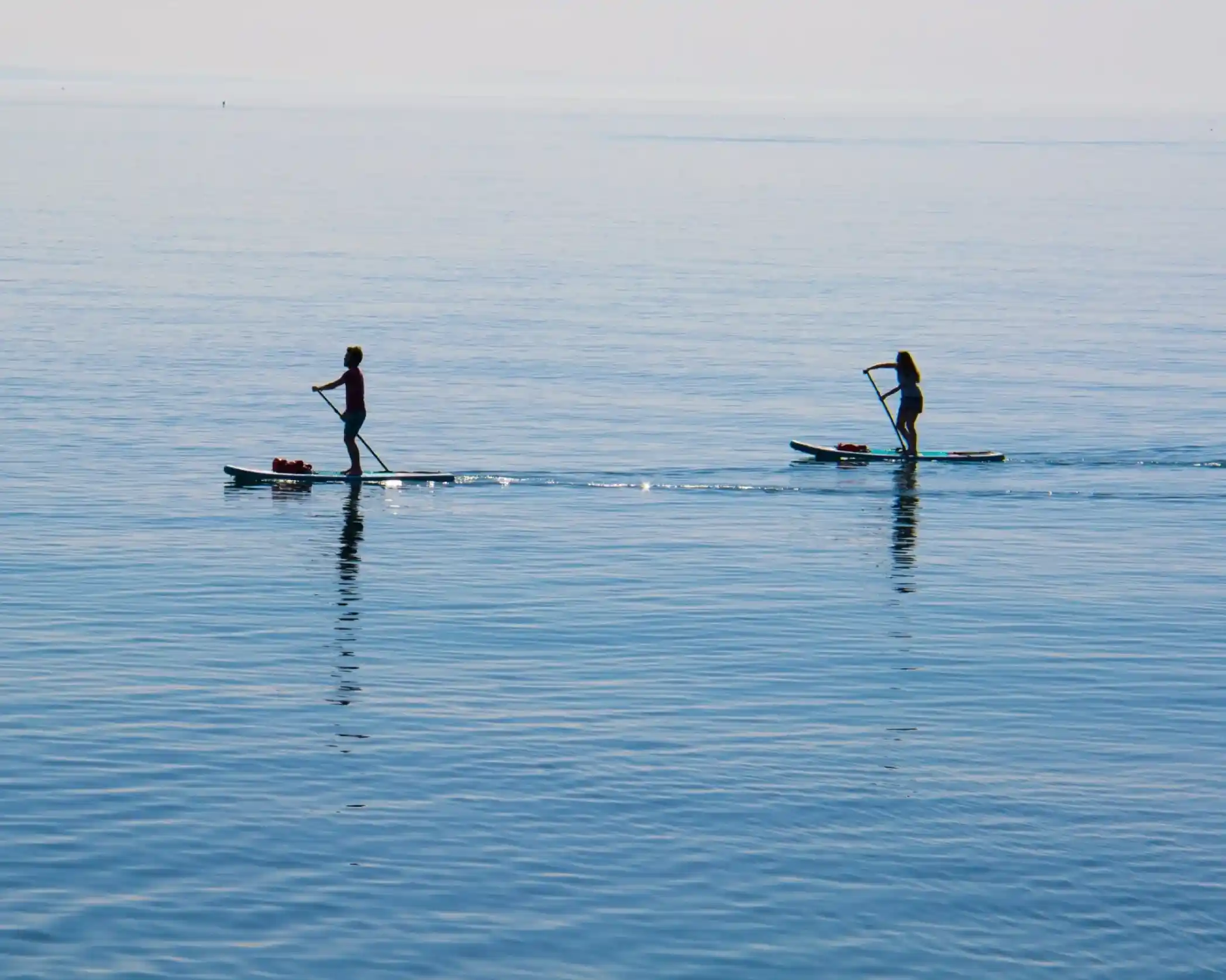 Two individuals paddleboarding on a calm, reflective lake with a serene blue surface.
