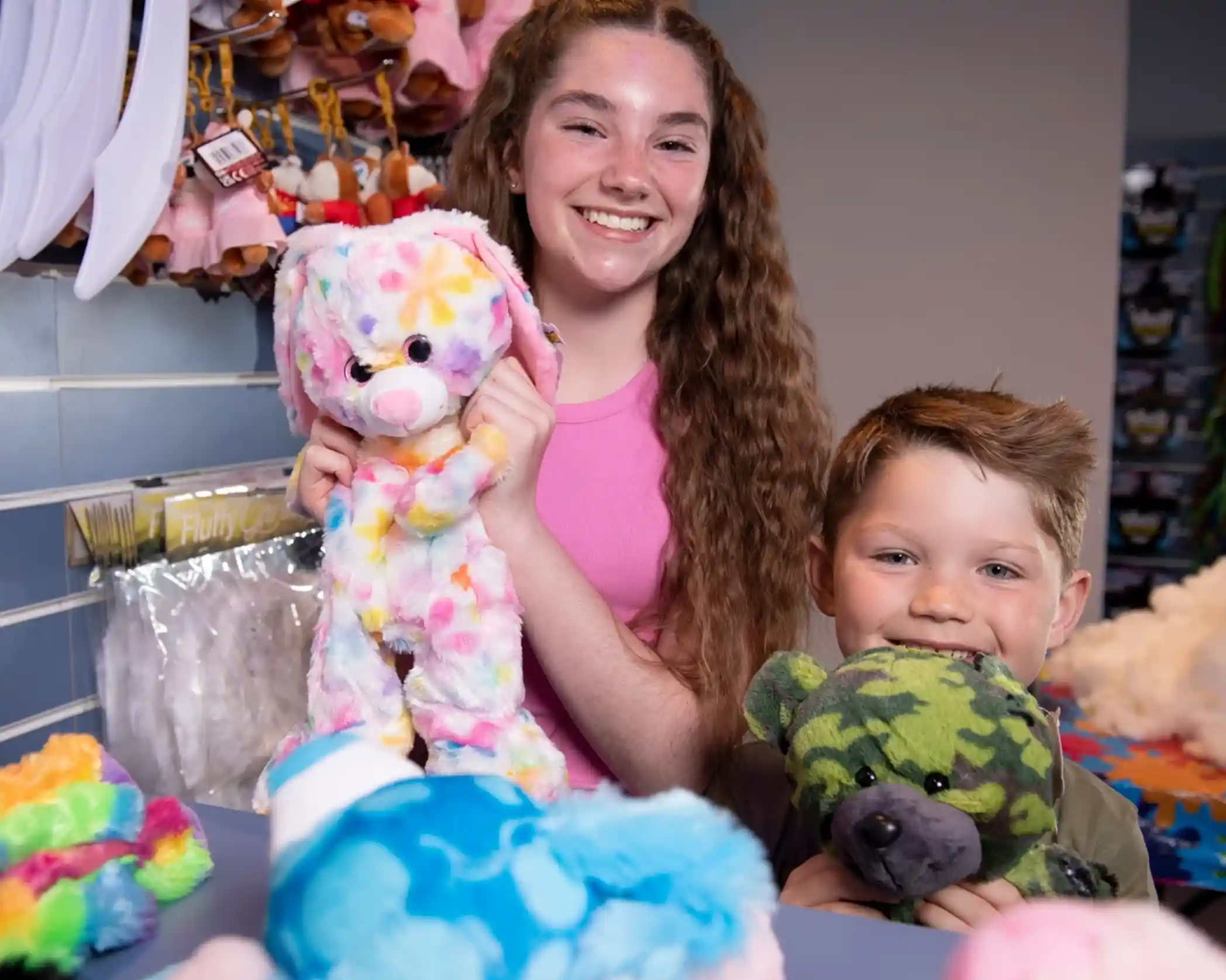 A teenage girl smiles while holding a colorful stuffed bunny, next to a young boy grinning with a green camouflage teddy bear. They are surrounded by various plush toys in a store.