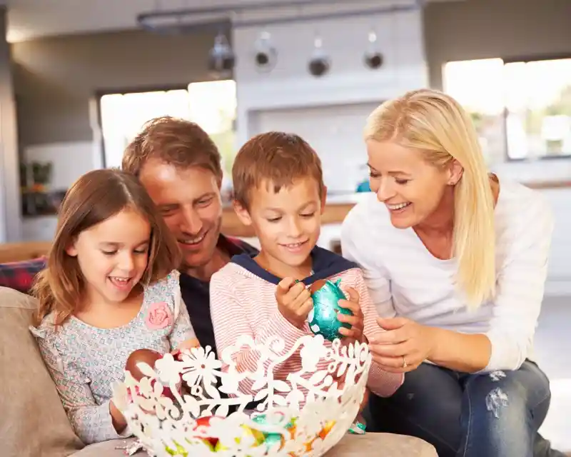 A family of four enjoys decorating colorful eggs together while sitting on a couch. Two children, a boy and a girl, are focused on their crafts, while the parents smile and assist them. The setting is bright and cozy, with an inviting kitchen visible in the background.