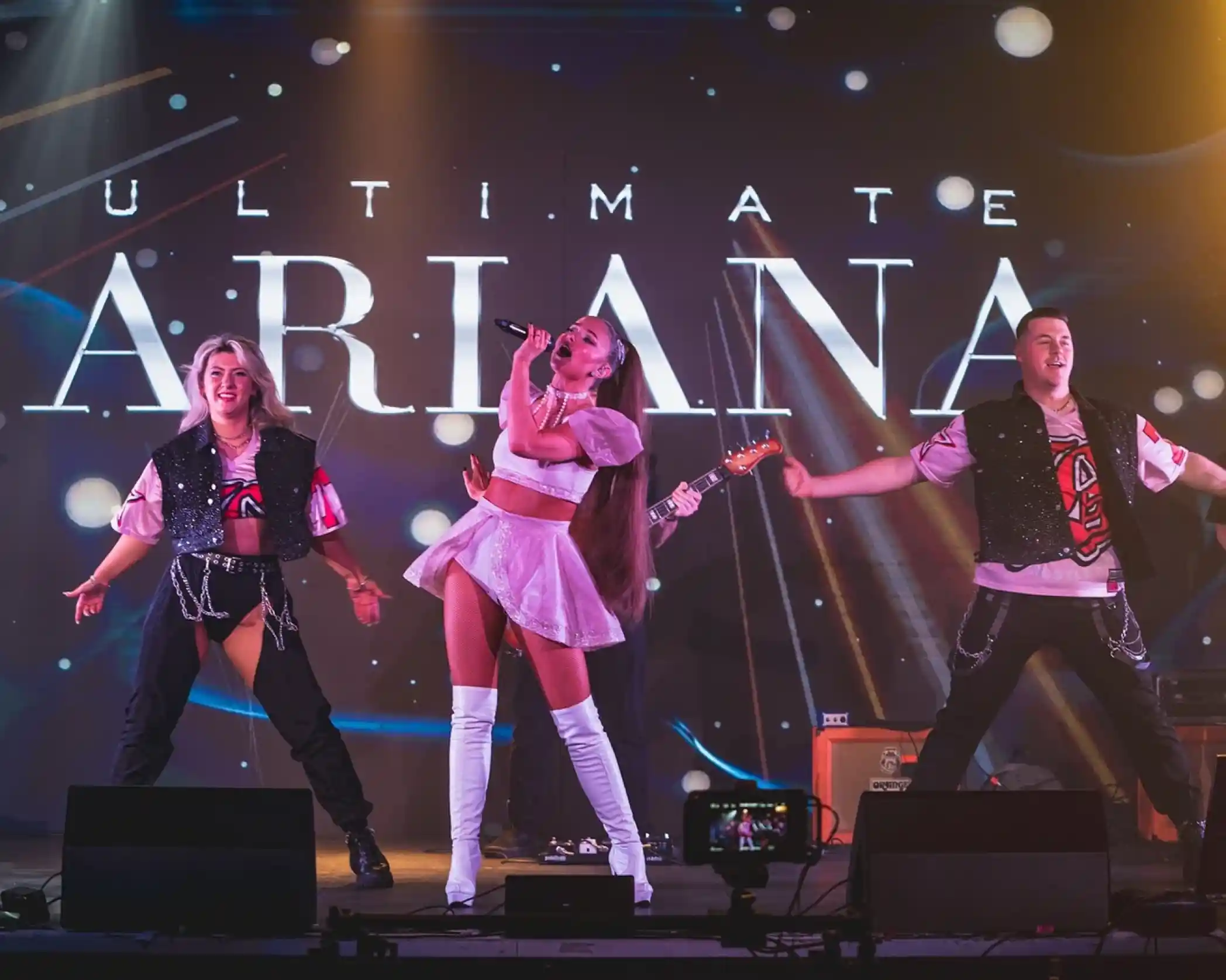 Performers on stage during a tribute show called "Ultimate Ariana," with one singer in a pink outfit and white boots, surrounded by two backup dancers. Colorful lights and a cosmic background enhance the vibrant atmosphere.
