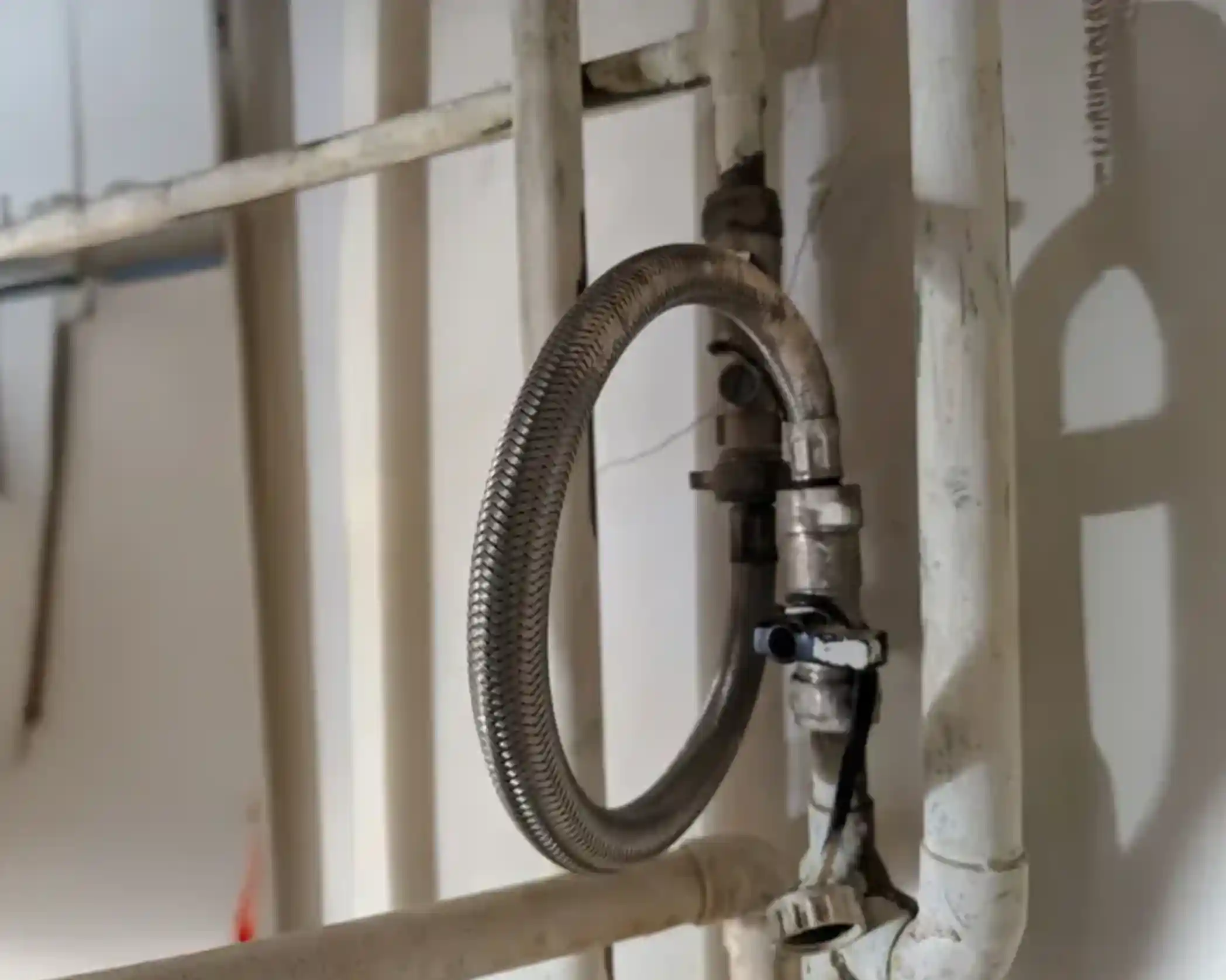 Close-up of metallic pipes and flexible hose connection on a wall. The pipes are painted white, and there is a small valve and a wiring connection visible.