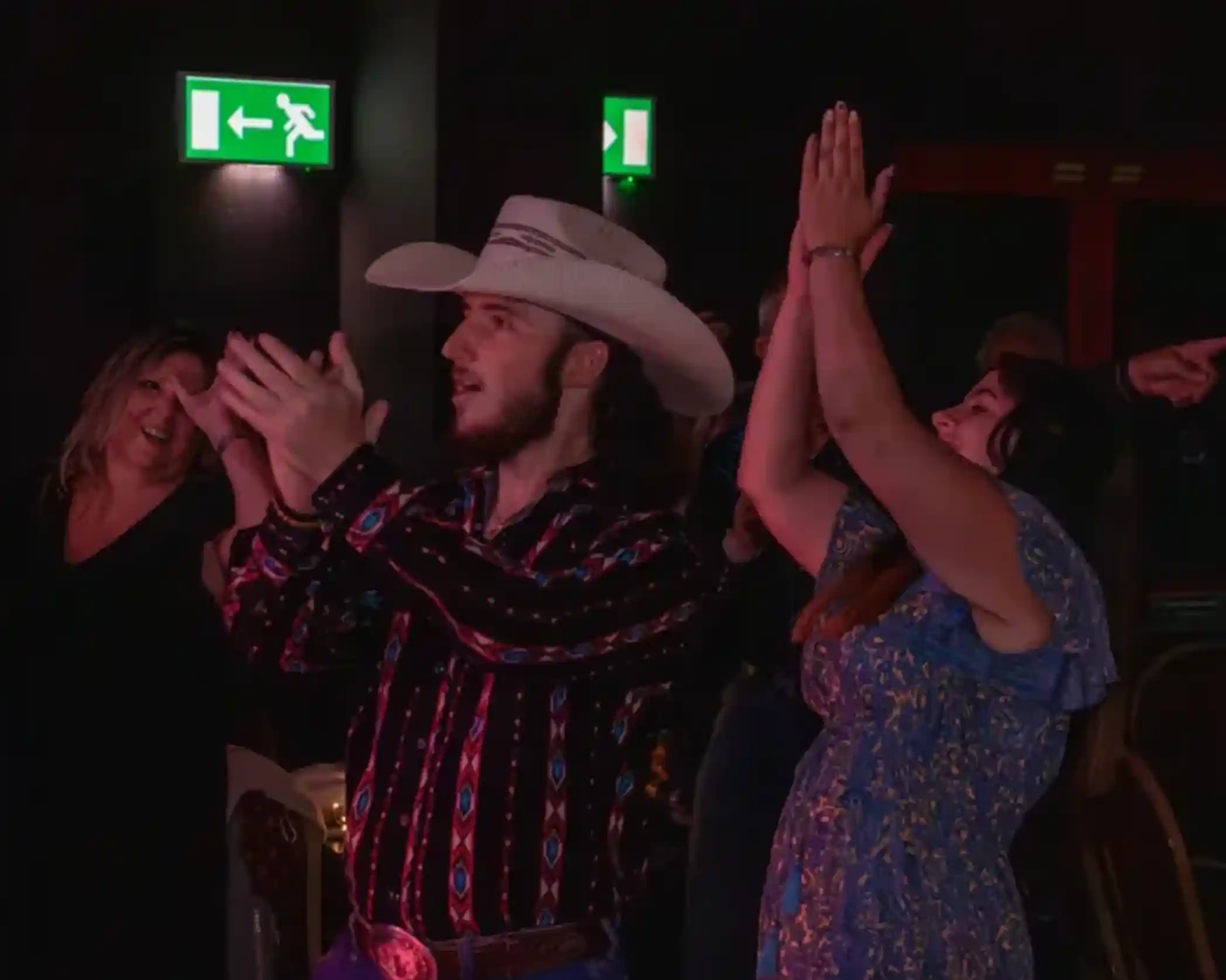 A group of people enjoying a night out, with a man in a cowboy hat and patterned shirt clapping his hands. A woman next to him is also clapping, and others are visible in the background celebrating. The scene is lit with dim lighting, creating a festive atmosphere.