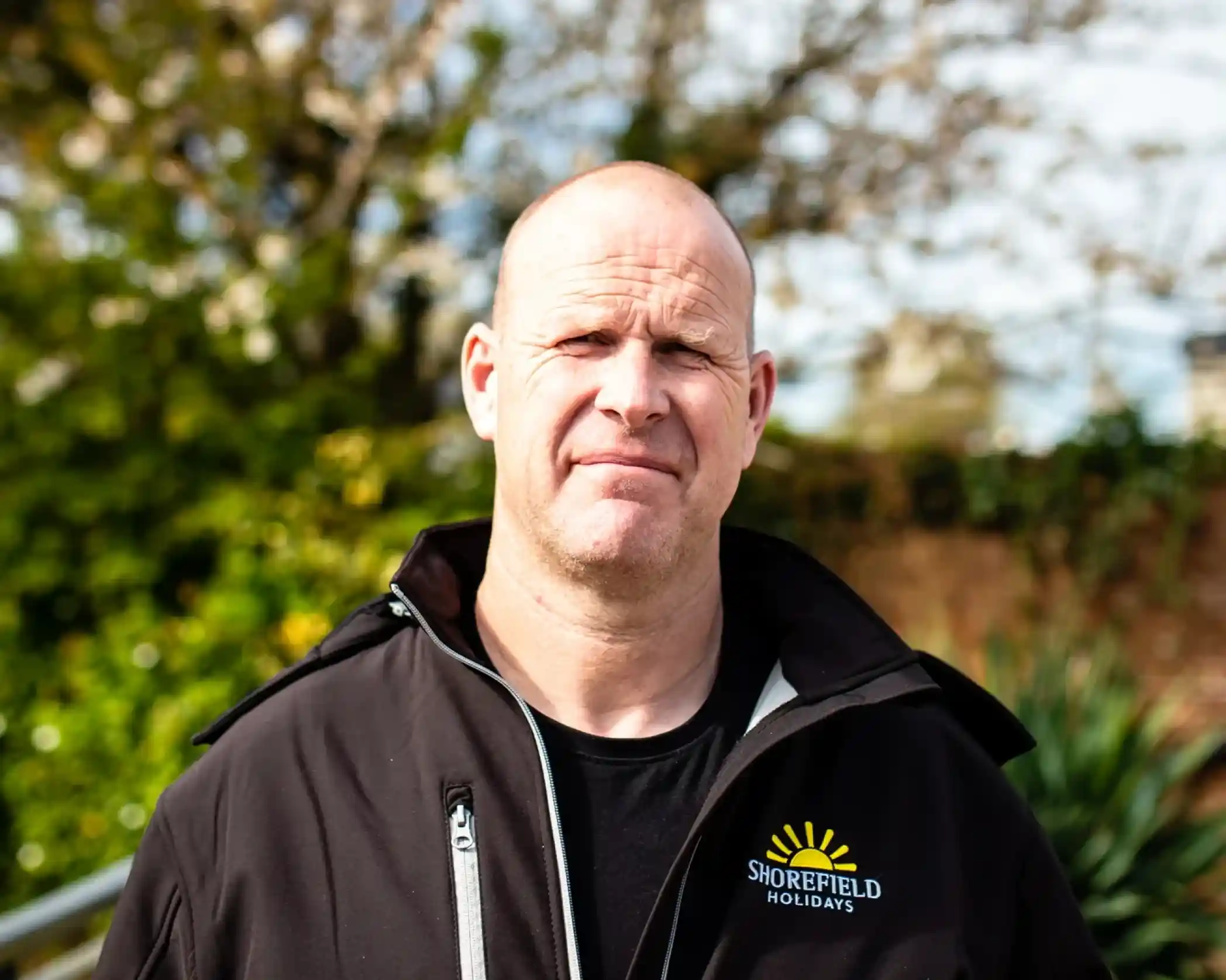 A man with a bald head is standing outdoors, wearing a black jacket with the logo of Shorefield Holidays. He appears to be looking directly at the camera, with greenery and blossoms in the background.