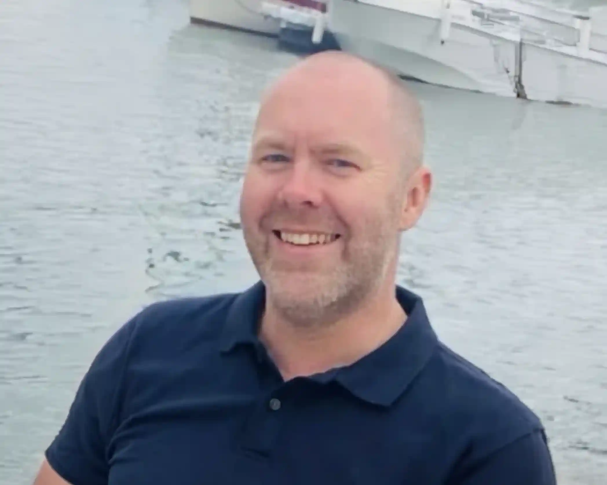 Smiling man with a short beard, wearing a navy polo shirt, sitting by the water with boats in the background.