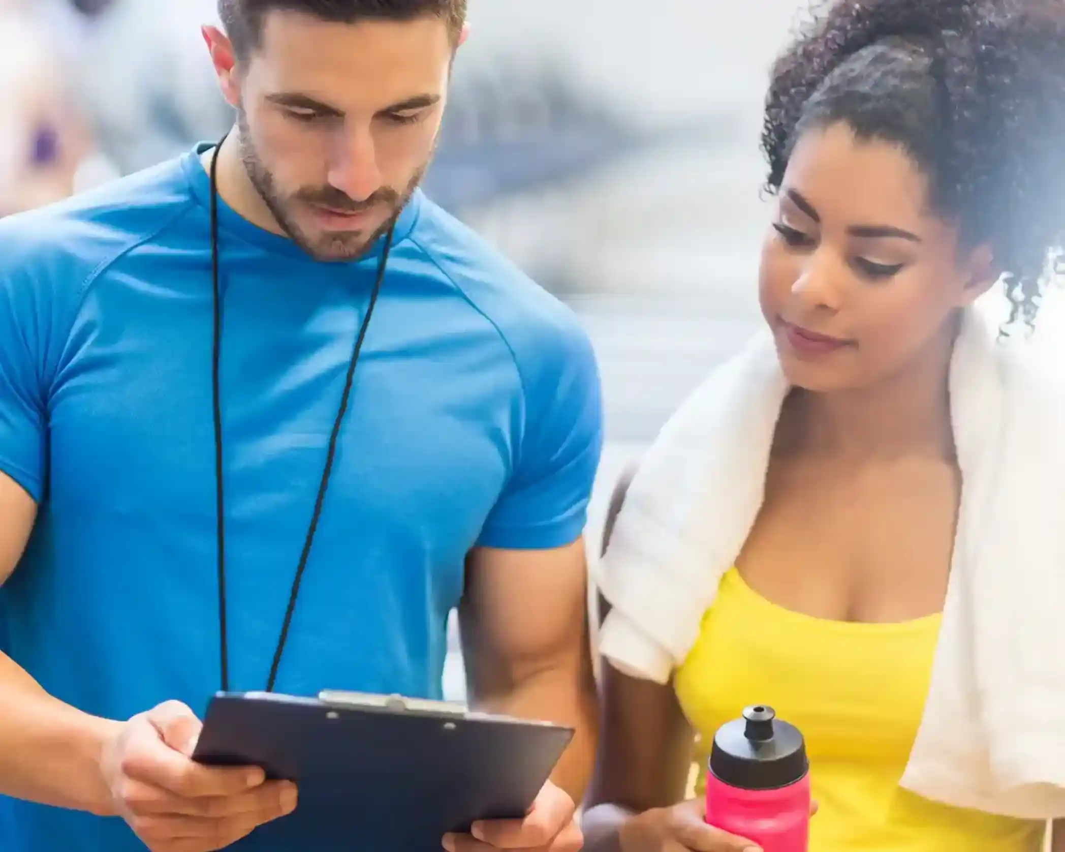 A fitness instructor and a woman are discussing information on a clipboard. The instructor is in a blue shirt, while the woman is wearing a yellow tank top, holding a water bottle. A towel is draped over her shoulder.