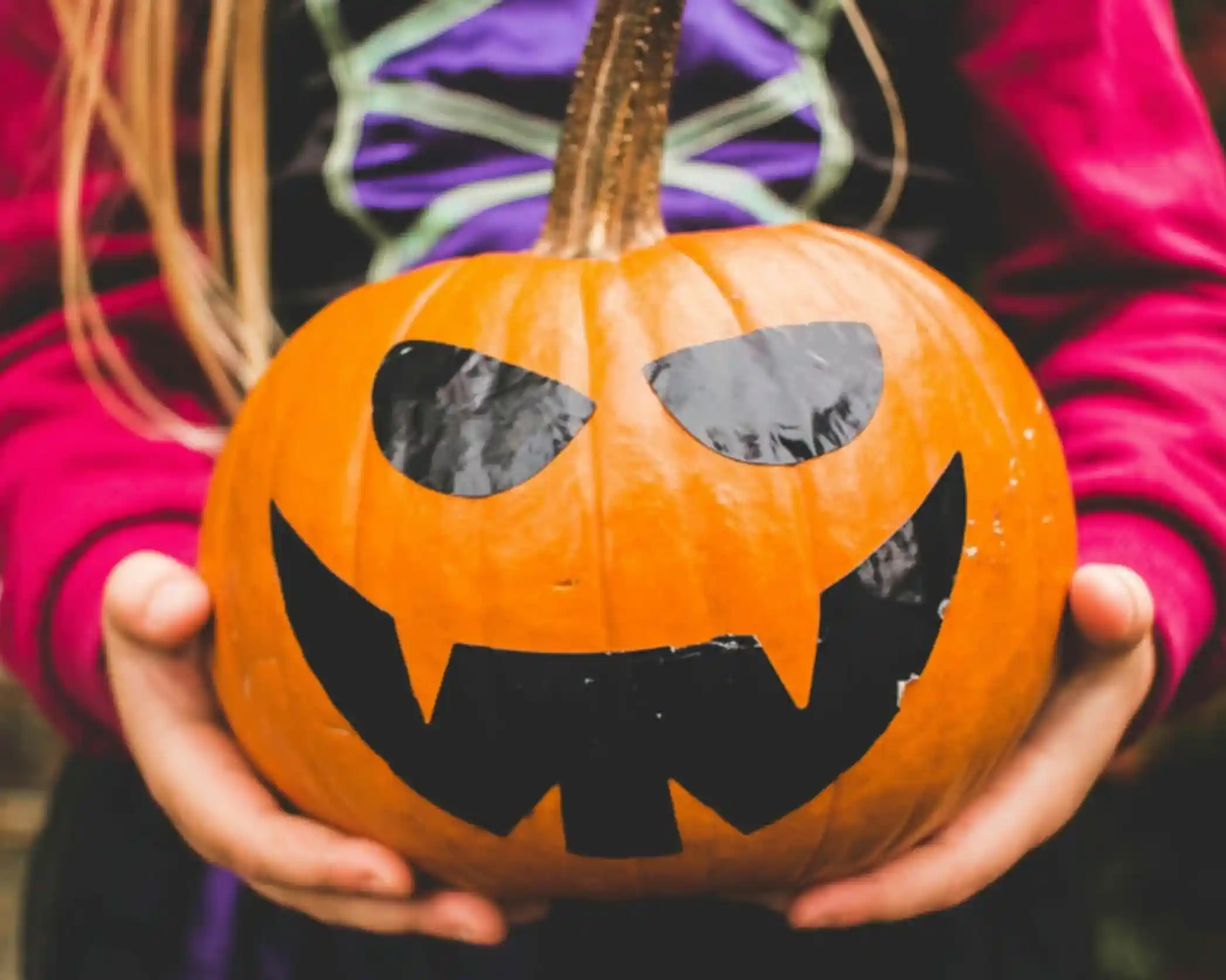 A person holds a carved pumpkin with a mischievous face, featuring black eyes and a toothy grin. The pumpkin has a vibrant orange color and a long stem, set against a blurred background.