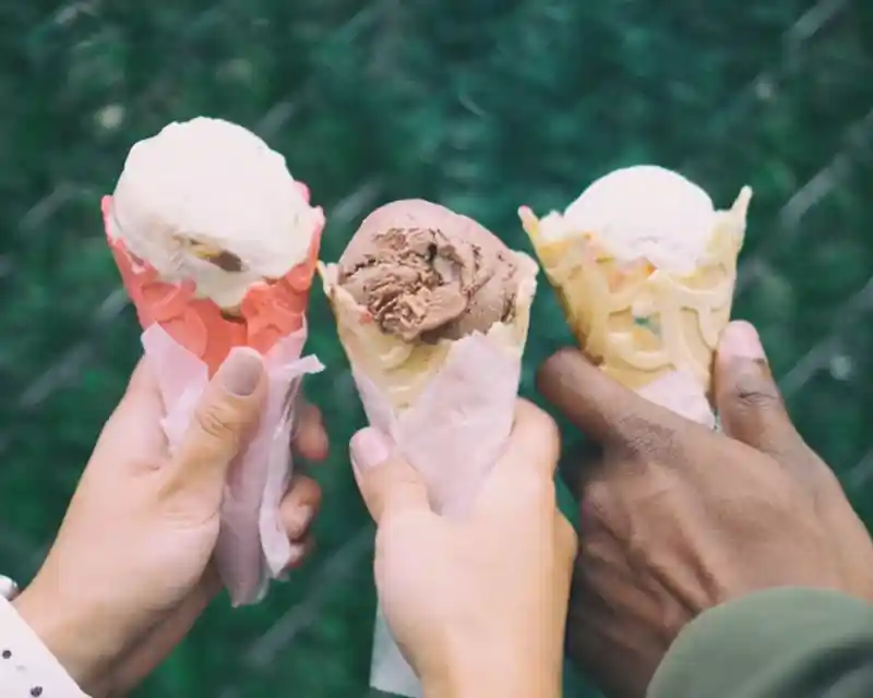 Three hands hold colorful ice cream cones, each topped with different flavors. The cones are wrapped in paper, set against a green background.