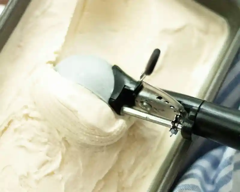 A close-up of an ice cream scoop dishing out a round scoop of vanilla ice cream from a metal container.