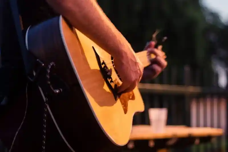 A close-up of a person's hand playing an acoustic guitar, with a blurred background featuring a table and greenery.