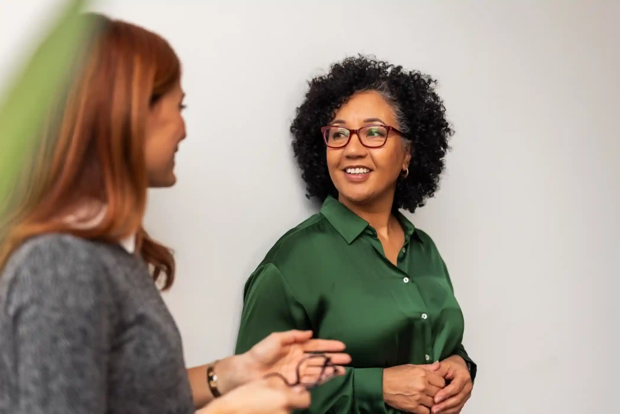 Two women engage in a conversation. One woman with curly hair and glasses wears a green blouse, while the other has straight red hair, dressed in a gray sweater. They appear to be smiling and enjoying each other's company against a neutral background.