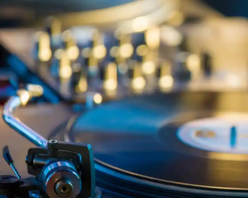 A close-up of a turntable with a spinning vinyl record and a stylus arm in focus. The background features blurred knobs and buttons, suggesting a DJ setup.