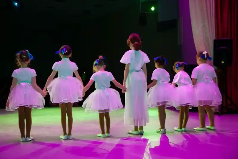 A group of seven young girls in white dresses holding hands, facing away from the camera. They stand on a stage with purple and green lighting, creating a festive atmosphere.