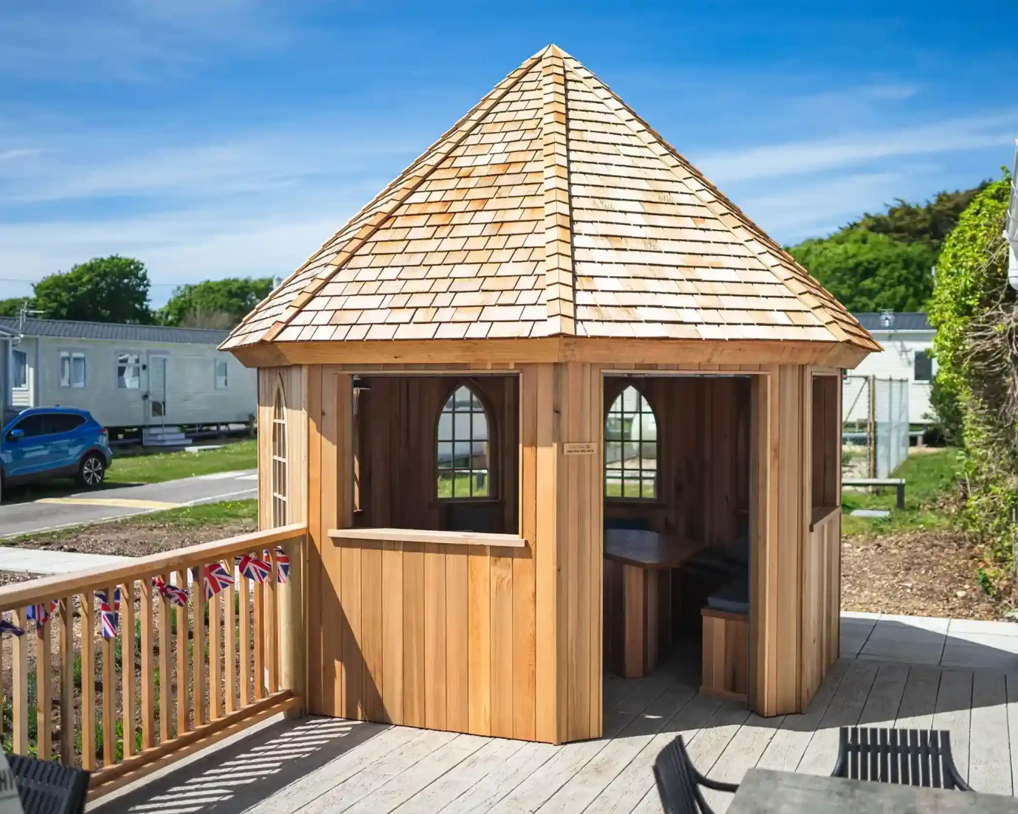 A wooden gazebo with a pointed shingle roof, featuring arched windows and a wraparound railing. It's situated on a deck overlooking a park-like setting with caravans in the background.