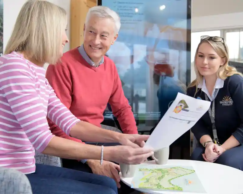 A man and two women are engaged in a discussion while sitting at a table. The man, dressed in a red sweater, is smiling as he looks at the women. One woman, wearing a striped shirt, holds a document, while the other, in a uniform, listens attentively. A colorful map and drinks are on the table.