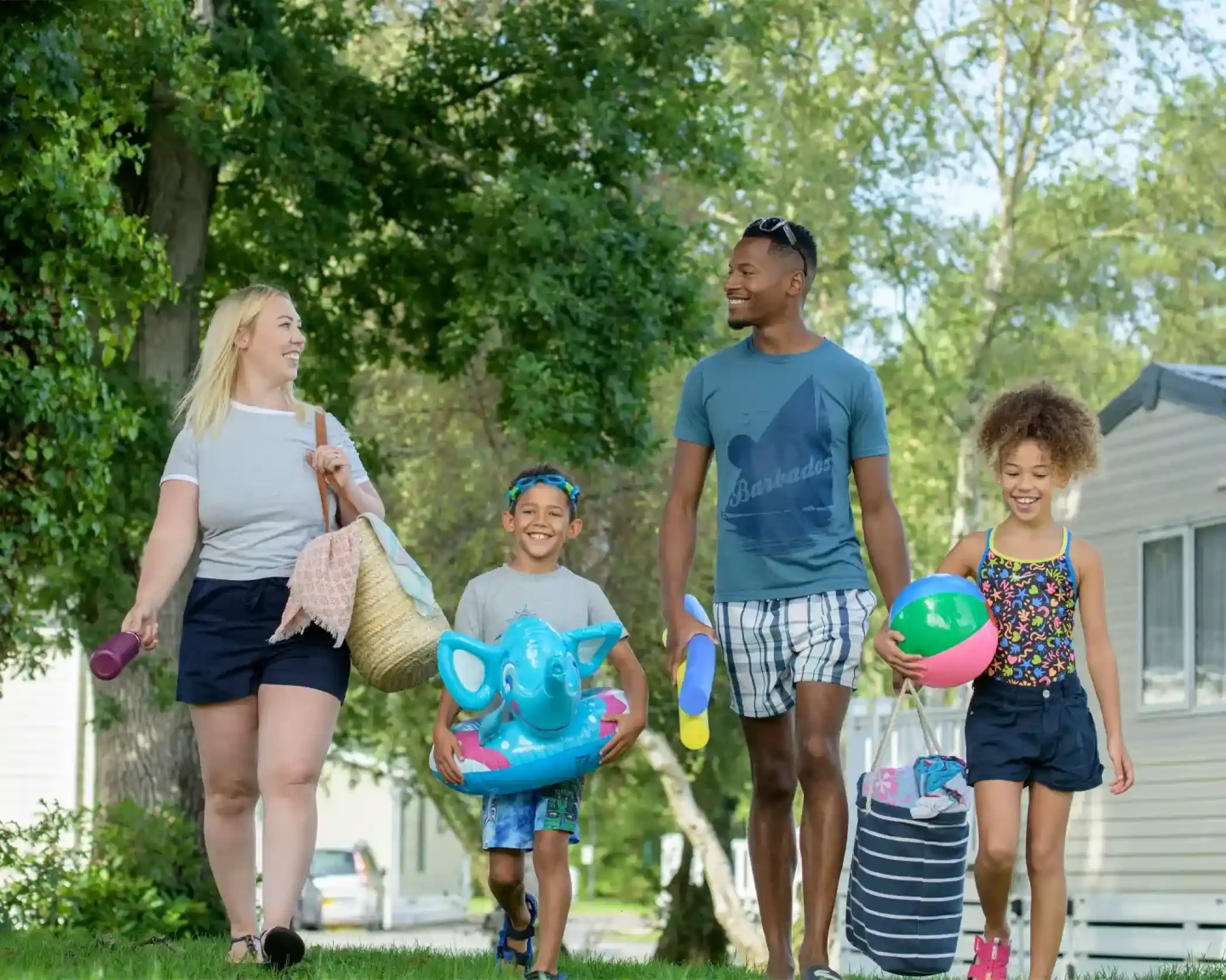 A family enjoys a sunny day while walking together. Two children, a boy and a girl, hold colorful beach toys, while an adult man and woman carry bags. They smile and chat as they stroll along a grassy path, surrounded by trees and mobile homes.
