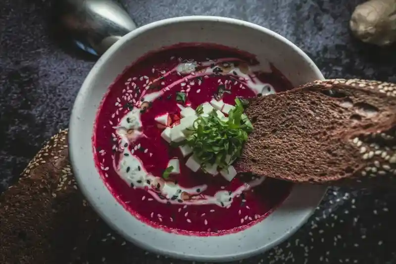 A bowl of vibrant beetroot soup topped with diced vegetables, chopped herbs, and drizzled with cream. A piece of dark bread is being dipped into the soup. The setting has a dark, textured background.
