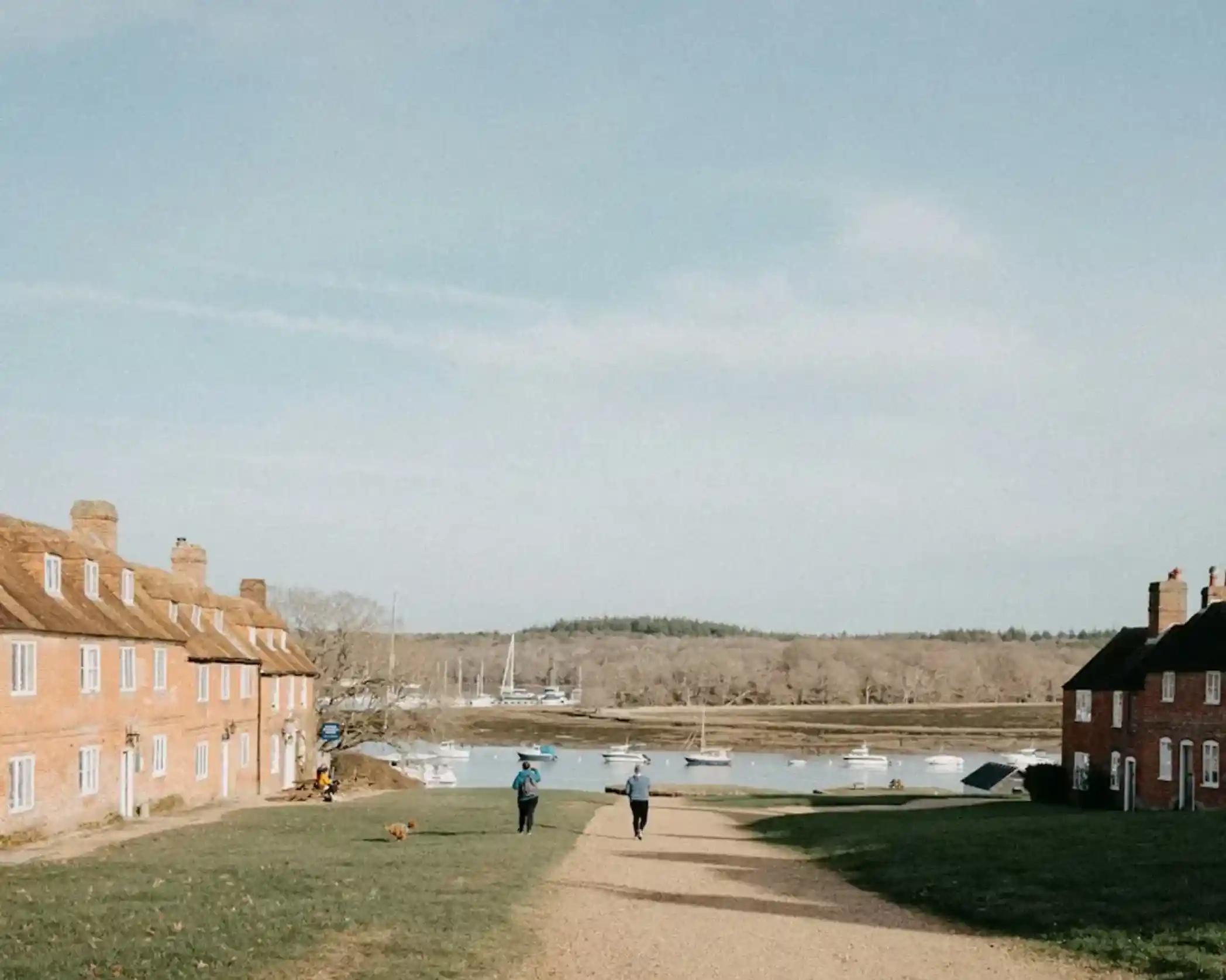 Two people walk down a gravel path lined with red-brick houses, with a view of a tranquil waterway and boats in the distance. The scene is set against a clear, blue sky.