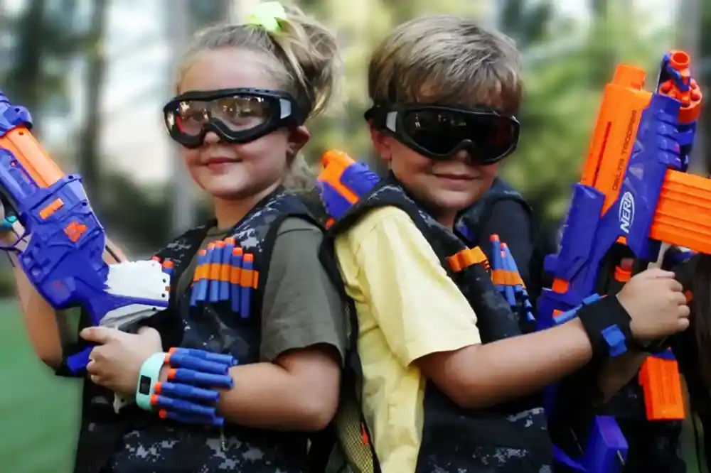 Two children stand back-to-back, wearing tactical vests with colorful toy dart guns. They both wear protective goggles and smile confidently at the camera. The background features greenery, suggesting an outdoor setting.