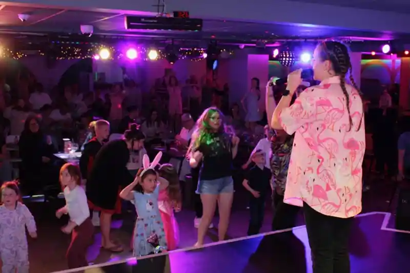 A performer with braided hair in a pink flamingo shirt sings on stage, while children and families dance in a colorful, lively venue with bright lights.