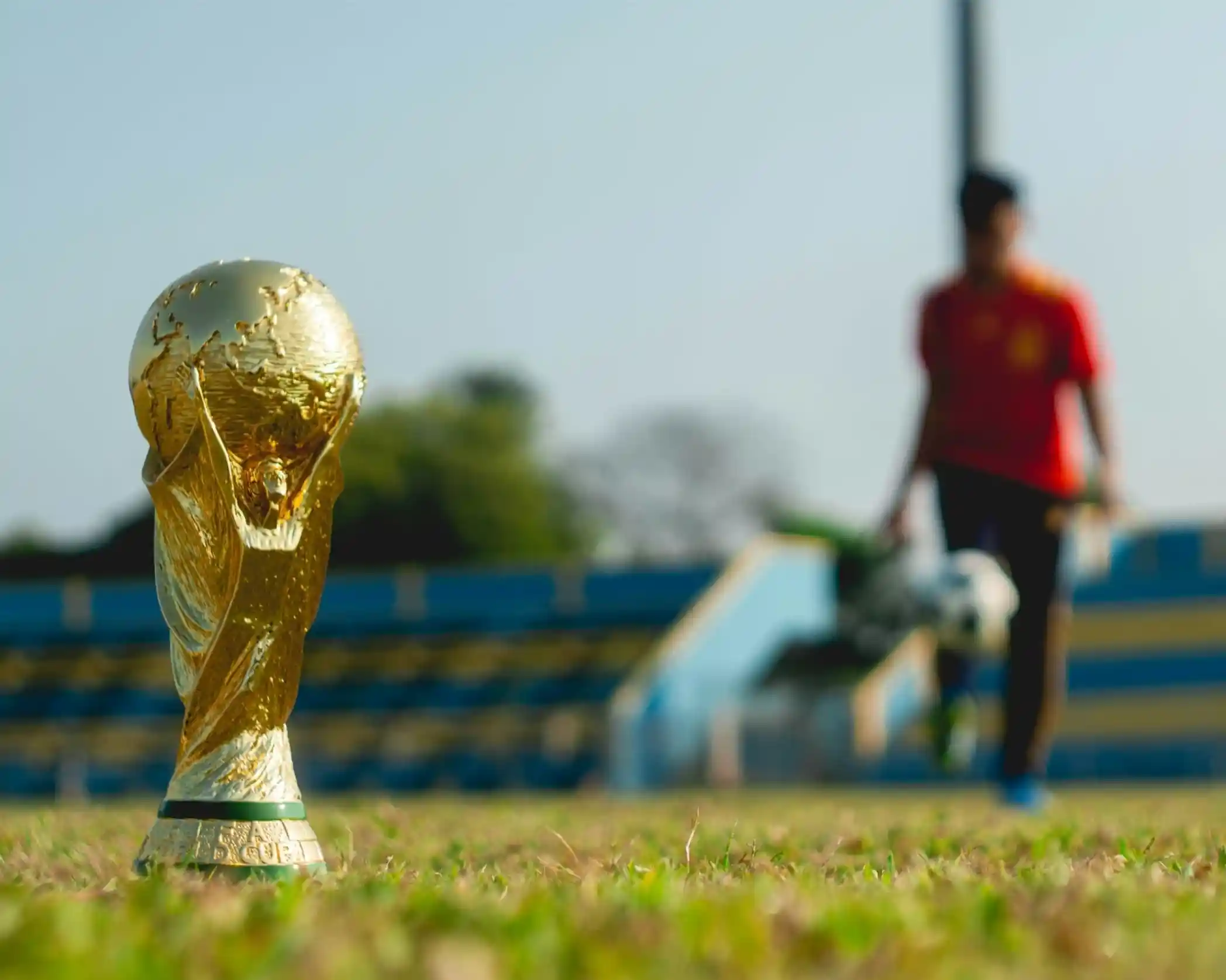 A golden World Cup trophy in the foreground with a blurred figure of a person holding a soccer ball in the background on a grassy field.