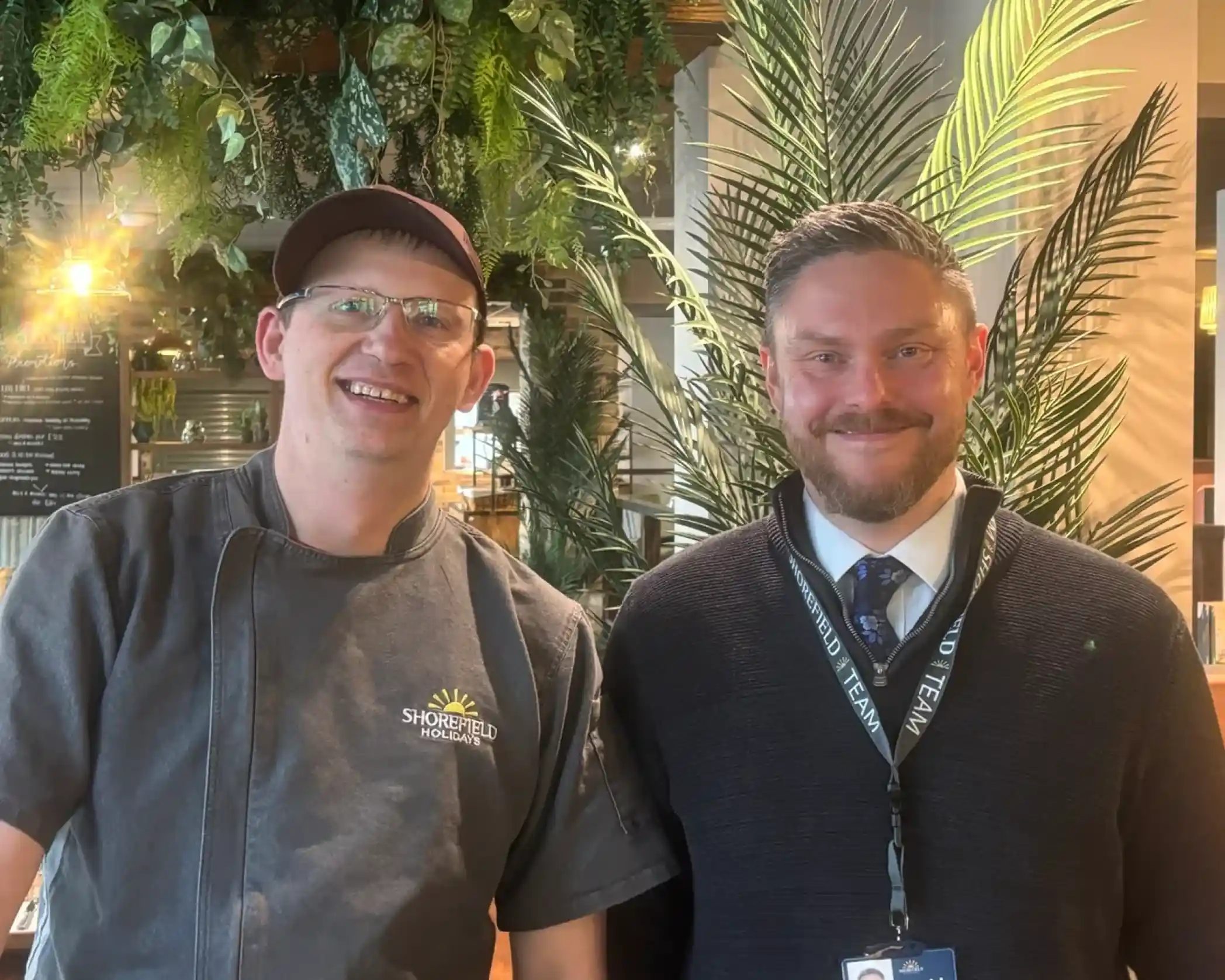 Two men stand smiling in a lush indoor setting with greenery in the background. One is wearing a chef's uniform and cap, while the other is dressed in a business suit with a lanyard.