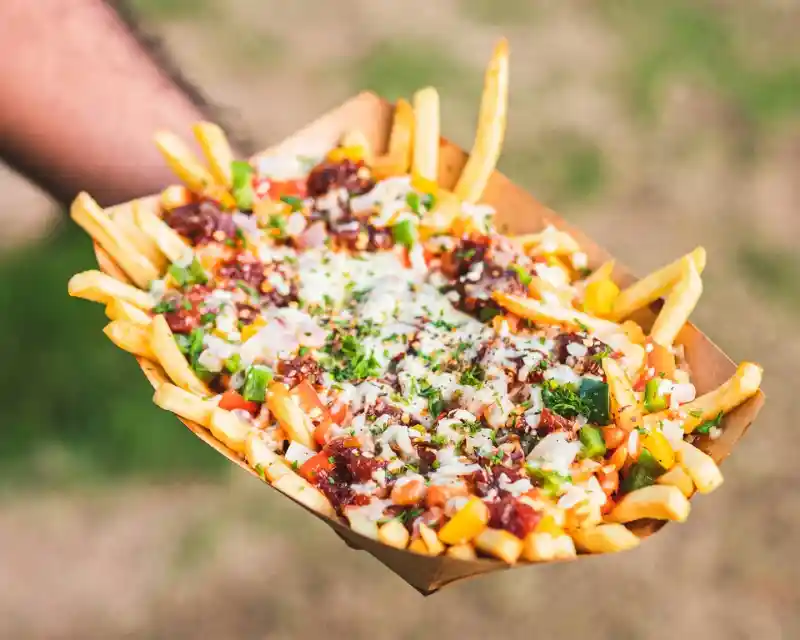 A hand holds a tray of loaded French fries topped with cheese, salsa, onions, and chopped herbs. The background is blurred greenery, enhancing the focus on the delicious dish.