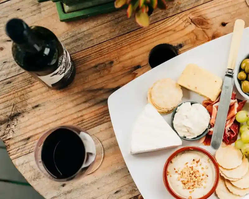 A white platter filled with various cheeses, olives, crackers, and dips sits on a wooden table. A glass of red wine and a bottle of wine are nearby, along with a small succulent plant.