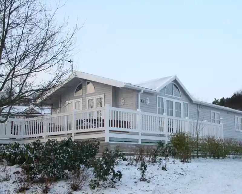A modern wooden cabin with a white deck surrounded by snow, featuring large windows and a sloped roof. Sparse vegetation is visible in the foreground, and a clear sky is seen above.