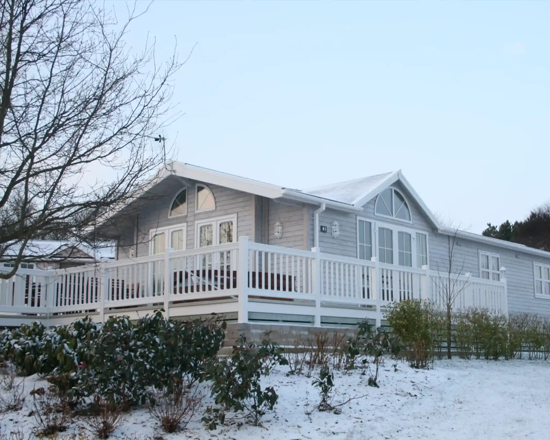 A modern wooden cabin with a white deck surrounded by snow, featuring large windows and a sloped roof. Sparse vegetation is visible in the foreground, and a clear sky is seen above.