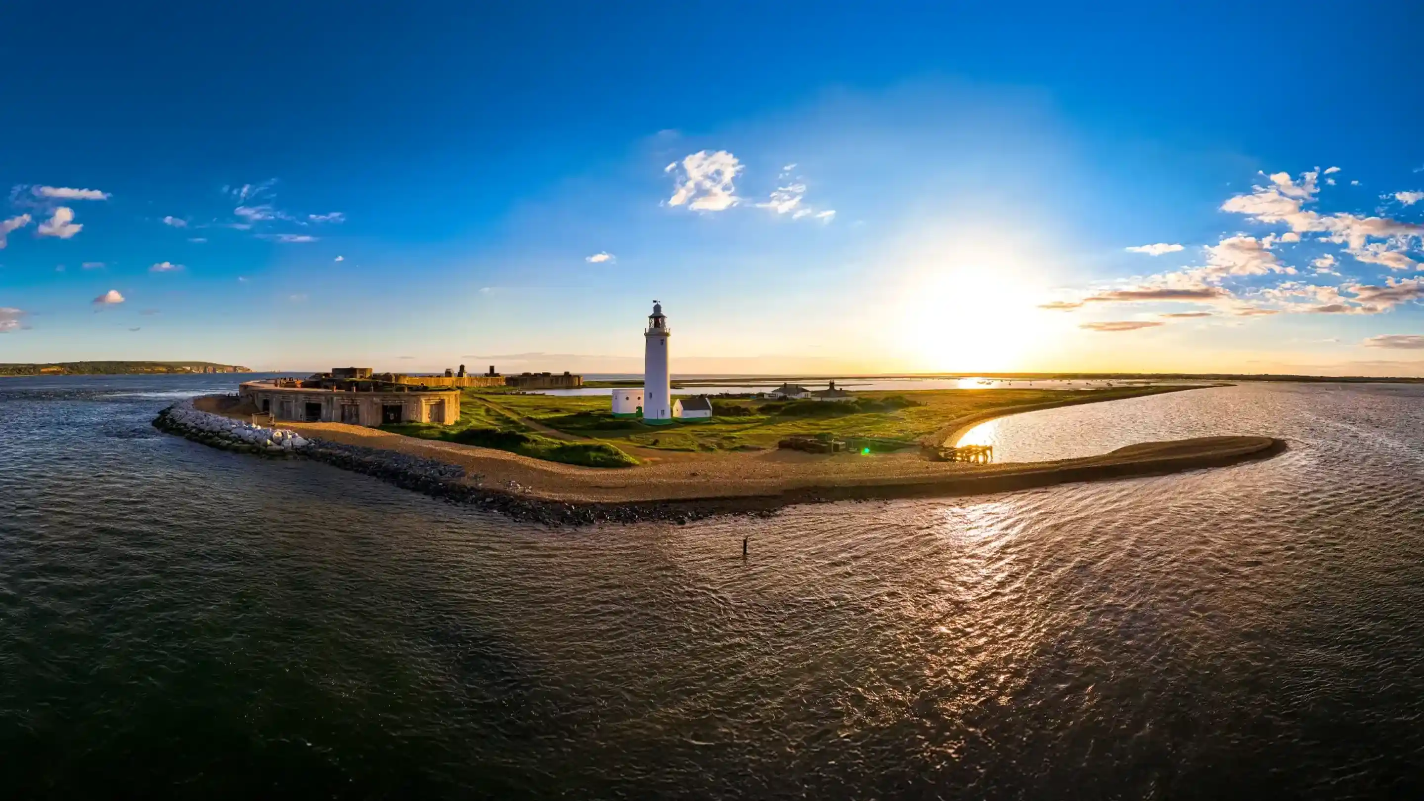 A scenic view of a lighthouse on a small island, surrounded by water, with a vibrant sunset in the background. The sky is clear with wispy clouds and the sunlight casts a warm glow over the landscape.
