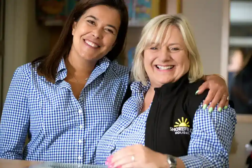Two smiling women stand together, both wearing checkered blue shirts. One woman has long dark hair, while the other has short blonde hair and wears a black vest with the logo "SHOREFIT HOLIDAYS." They appear friendly and welcoming.