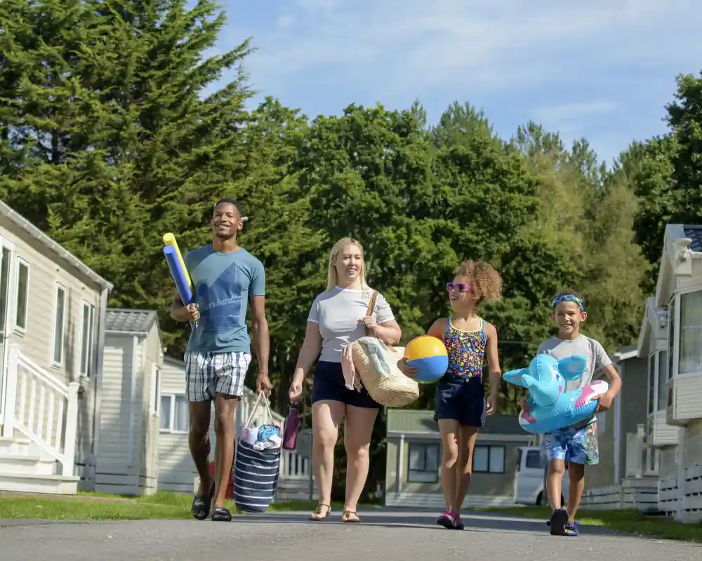 A family walks along a pathway lined with holiday homes. They carry beach toys, towels, and other supplies, with greenery in the background and a clear blue sky above.