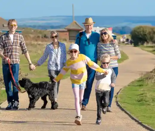 A cheerful family walks along a sunny path, holding hands and enjoying the outdoors. Two children run ahead, while adults smile and walk with a dog on a leash. Lush greenery and blue skies enhance the joyful scene.